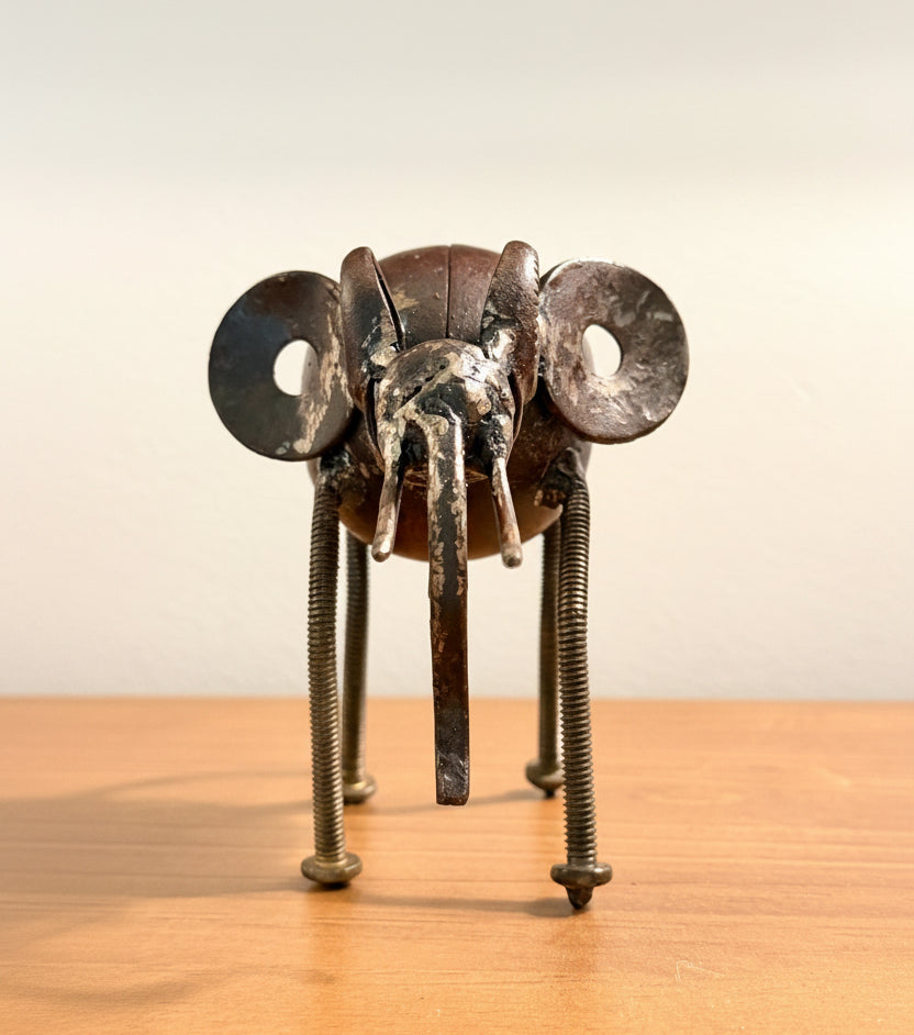 A rustic metal insect sculpture, resembling a beetle or grasshopper, crafted from scrap metal parts including circular side plates, a central body, and long threaded legs, lowered trunk frontal view