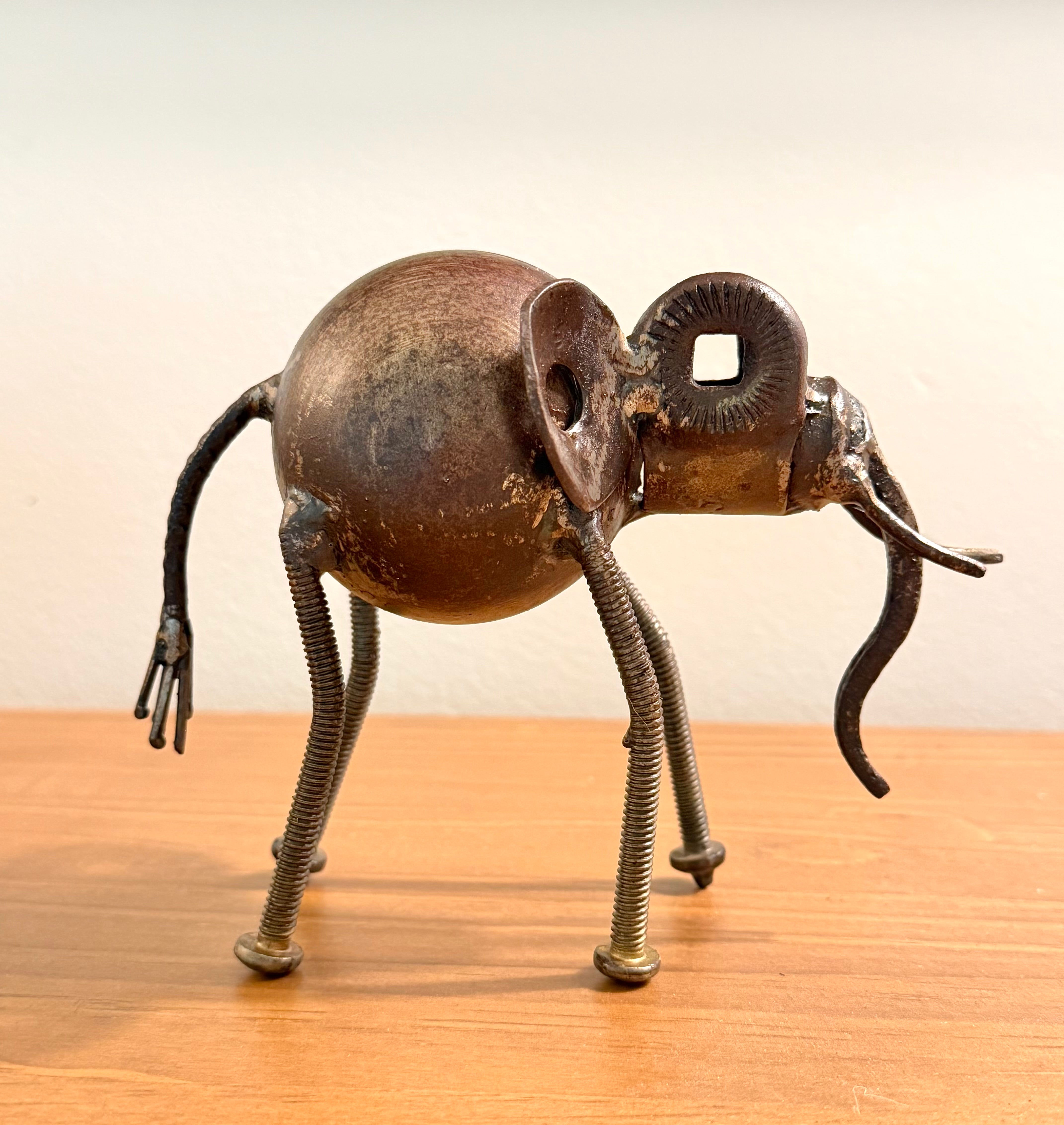 A rustic metal elephant figurine crafted from scrap materials, featuring a spherical body, twisted spring legs, and a curled lowered trunk
